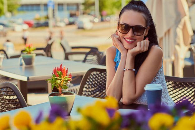 Das Porträt eines glücklichen, charmanten, brünetten Mädchens, das trendige Kleidung trägt, genießt den Sommertag, während es auf einer Terrasse im Café im Freien sitzt und in die Kamera schaut.