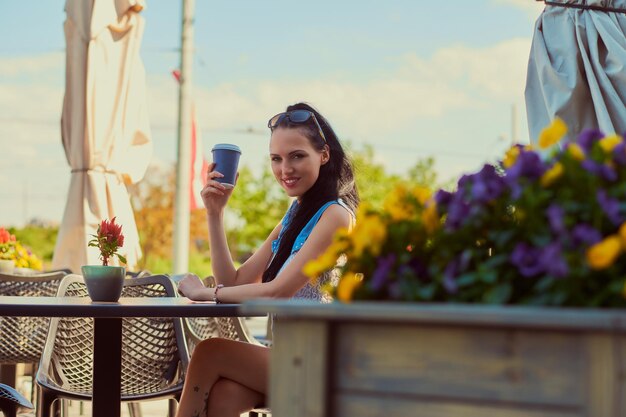 Das Porträt eines glücklichen, schönen Mädchens, das trendige Kleidung trägt, hält einen Kaffee zum Mitnehmen und genießt den Sommertag, während er auf einer Terrasse im Café im Freien sitzt.