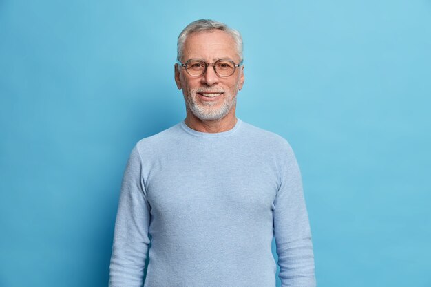 Das Porträt eines gutaussehenden bärtigen europäischen Mannes mit grauem Haar und Bartlächeln sieht angenehm direkt nach vorne aus, wenn es gut gelaunt ist, trägt der glückliche Tag eine Brille und einen Pullover, die über der blauen Wand isoliert sind