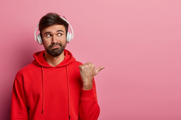 Das Porträt eines neugierigen bärtigen Mannes zeigt mit dem Daumen auf die leere Stelle rechts, trägt ein Stereo-Headset und ein rotes lässiges Sweatshirt und zeigt etwas Interessantes, das auf einer rosa Pastellwand isoliert ist.