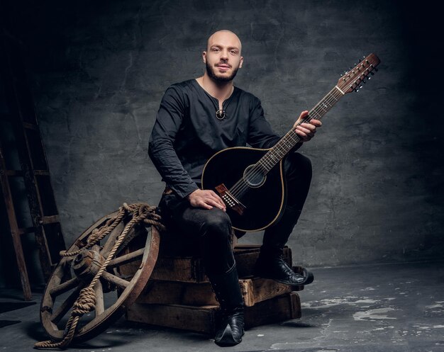 Das Porträt eines rasierten männlichen Musikers in alten keltischen Kleidern sitzt auf einer Holzkiste und spielt eine alte Mandoline.