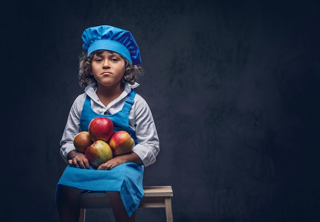 Das Porträt eines süßen kleinen Jungen mit braunen, lockigen Haaren, gekleidet in eine blaue Kochuniform, hält Äpfel, die auf einem Holzhocker in einem Studio sitzen. Getrennt auf dem dunklen strukturierten Hintergrund.