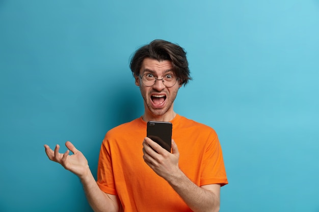 Das Porträt eines unzufriedenen wütenden erwachsenen Mannes hat eine verwirrte Reaktion auf das Lesen negativer Nachrichten über Mobiltelefone, Ausrufe und Gesten, hält ein Handy in der Hand, trägt Freizeitkleidung und posiert an der blauen Wand