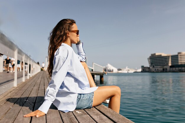 Das Profilporträt eines stilvollen europäischen Mädchens in blauem Hemd und Jeansshorts sitzt auf einem Holzsteg und freut sich im Sonnenlicht auf den Hintergrund eines blauen Sees mit Yachten