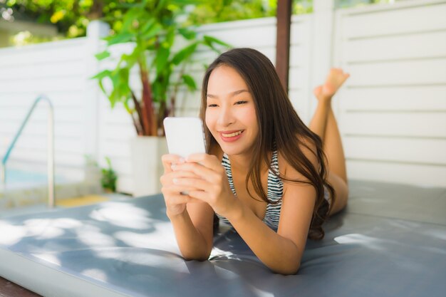 Das schöne junge asiatische Frauenlächeln des Porträts, das glücklich ist, entspannen sich mit Handy um Swimmingpool