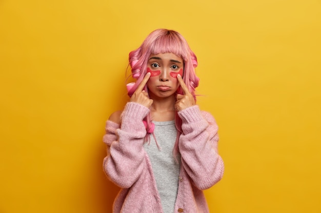 Das Schönheitsporträt des traurigen Mädchens zeigt auf kosmetische Kollagenflecken unter den Augen, hat düsteren Ausdruck, hat rosa Haare mit Fransen, trägt Lockenwickler