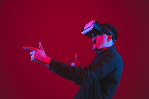 Das Spielen trug VR-Headset. Porträt des kaukasischen jungen Mannes isoliert auf roter Wand im Neonlicht. Schönes Modell. Konzept der menschlichen Emotionen, Gesichtsausdruck, Jugend, Geräte.