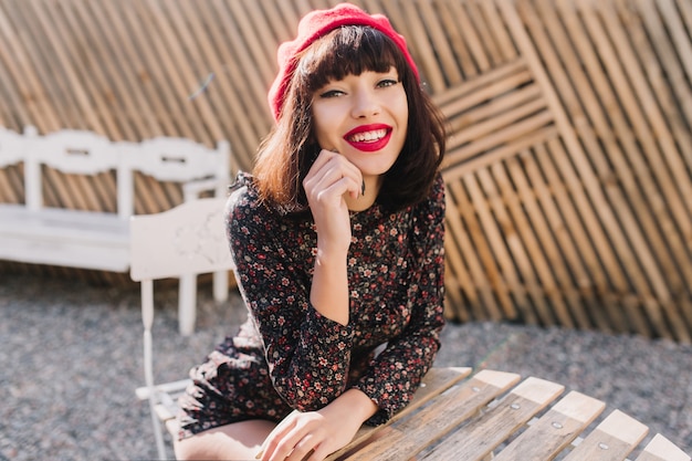 Das stilvolle brünette französische Mädchen in der trendigen roten Baskenmütze mit hellem Make-up kam zum Mittagessen ins Straßencafé. Porträt der eleganten jungen Frau in den Vintage-Kleidern, die auf ihre Bestellung im Freiluftrestaurant warten.