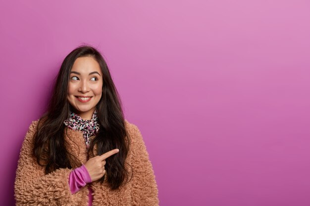 Das Studiobild einer positiven asiatischen Dame zeigt auf den Kopierbereich und empfiehlt, den Laden zu besuchen, in dem sie neue Kleidung für den Winter gekauft hat