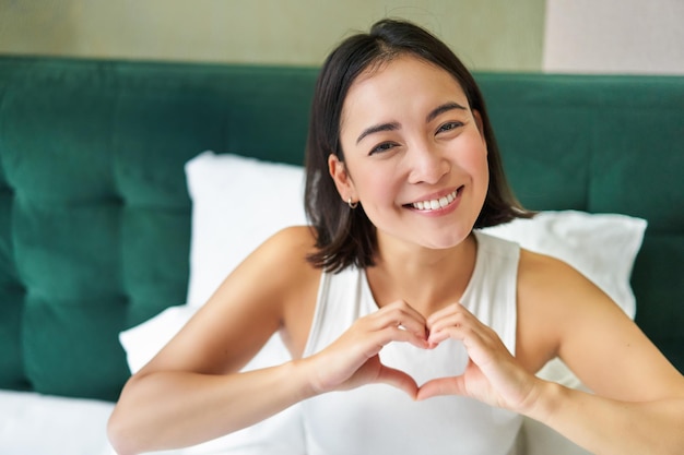 Das süße asiatische Mädchen zeigt ein Herzliebeszeichen, das im Bett liegt und lächelt und morgens faule Tage im Schlafzimmer genießt