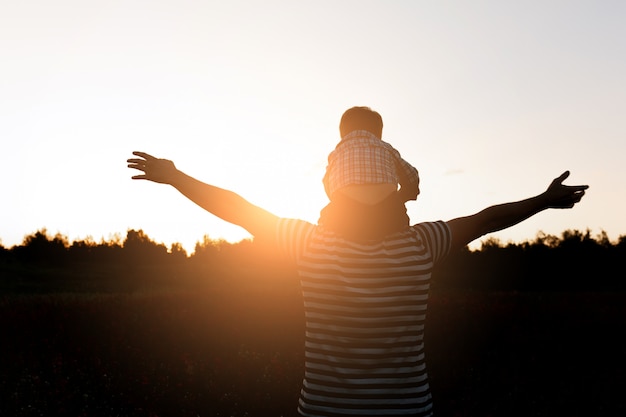 Das Vater- und Sohnschattenbild, das auf das Feld zur Sonnenuntergangzeit geht, der Junge, der sitzt, bemannt Schultern. Co