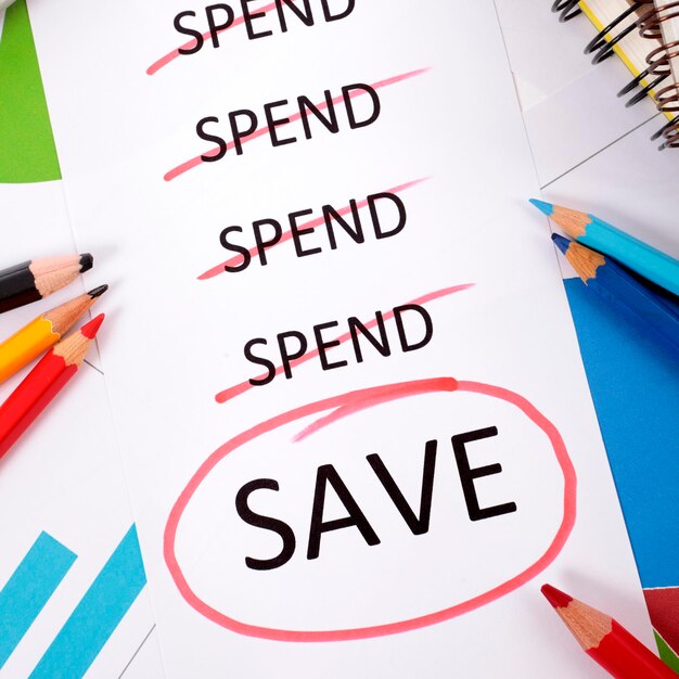 Das Wort "Save" ist rot eingekreist unter einer Ausgabenliste, die von Bleistiften, Grafiken, Büchern und Taschenrechnern umgeben ist.