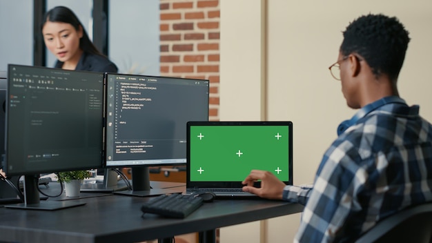 Datenbankdesigner, der Code mit einem Laptop mit Greenscreen-Chroma-Key-Mockup schreibt, der am Schreibtisch in einer geschäftigen Entwickleragentur sitzt. Programmierer, der mehrere Bildschirme mit Programmiersprache betrachtet.