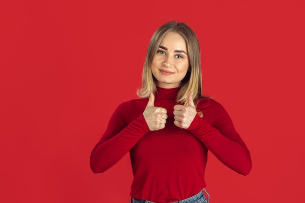 Kostenloses Foto daumen hoch. monochromes porträt der jungen kaukasischen blonden frau lokalisiert auf roter wand. schönes weibliches modell im hemd. menschliche emotionen, gesichtsausdruck, verkauf, anzeigenkonzept. jugendkultur.