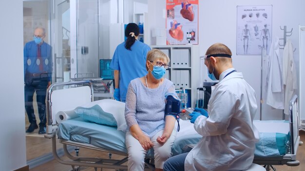 Dcotor überprüft Herzprobleme der alten Rentnerin während der Pandemie in einer modernen Privatklinik oder einem Krankenhaus. Patienten und medizinisches Personal tragen Masken zum Schutz vor COVID-19. Medizinisches Gesundheitswesen