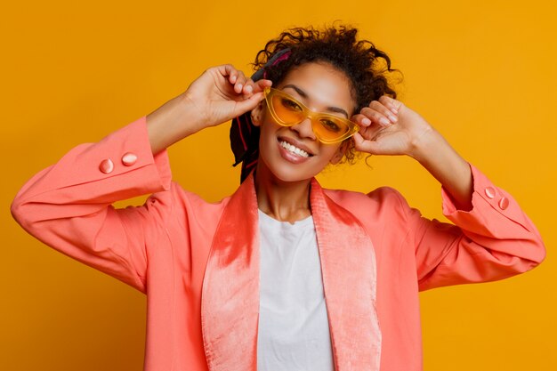 Debonair-Afrikanerin mit perfektem Lächeln, lockigen Haaren und natürlichem Make-up, das in der rosa trendigen Jacke auf gelbem Hintergrund im Studio aufwirft.