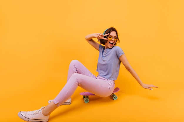 Debonair kaukasisches Mädchen trägt weiße Schuhe, die auf Skateboard sitzen. Lachende hübsche Dame in Kopfhörern posiert.