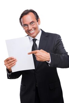 Dein text hier! glücklicher reifer mann in formeller kleidung, der leeres papier hält und mit einem lächeln darauf zeigt, während er isoliert auf weißem hintergrund steht