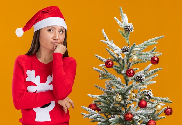 Denken, das junges asiatisches Mädchen der Seite betrachtet, das Weihnachtshut mit Pullover trägt, der nahe Weihnachtsbaum steht, der Hand auf Kinn lokalisiert auf orange Hintergrund setzt