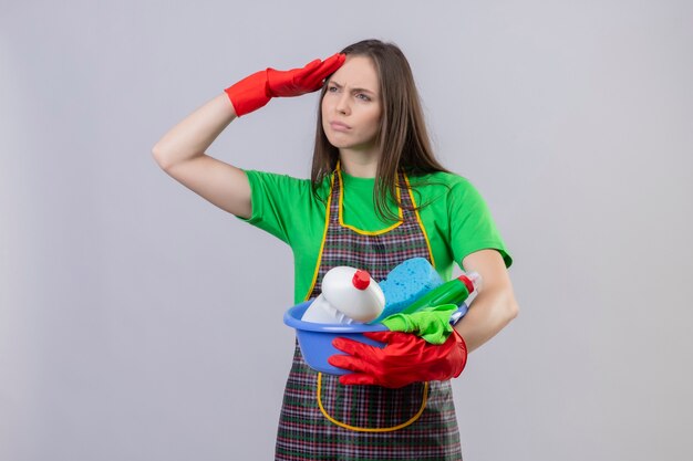 Denken, dass das Reinigen des jungen Mädchens, das Uniform in den roten Handschuhen hält, die Reinigungswerkzeuge halten, distany mit Hand auf isolierter weißer Wand betrachten