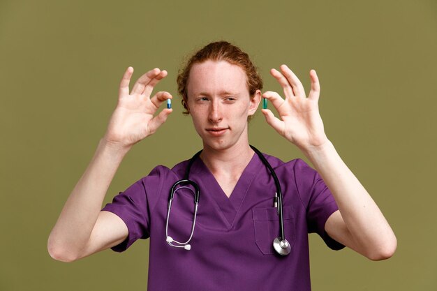 Denken halten Pillen junger männlicher Arzt in Uniform mit Stethoskop isoliert auf grünem Hintergrund