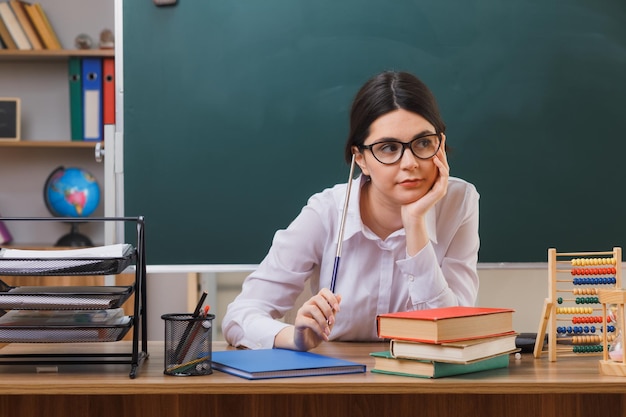 denken Hand aufs Kinn legen junge Lehrerin mit Brille und Zeiger am Schreibtisch sitzend mit Schulwerkzeugen im Klassenzimmer