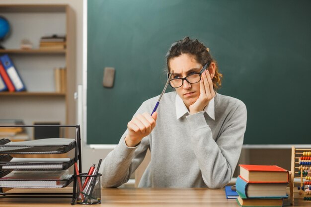 Denken, Hand aufs Kinn legen junger männlicher Lehrer, der eine Brille trägt und einen Zeiger hält, der am Schreibtisch mit Schulwerkzeugen im Klassenzimmer sitzt