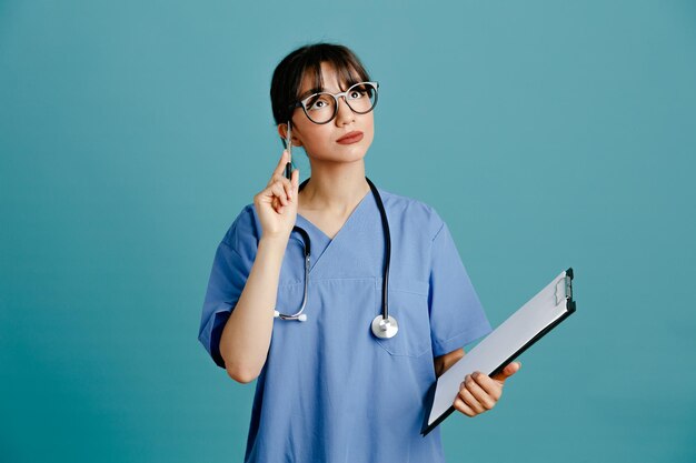 Denken Holding Klemmbrett mit Stift junge Ärztin mit einheitlichem fith Stethoskop isoliert auf blauem Hintergrund