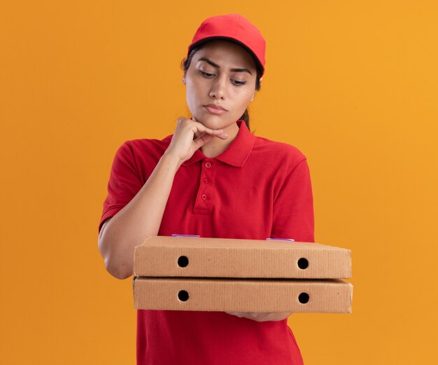 Denken junges Liefermädchen, das Uniform und Kappe hält und Pizzakästen betrachtet, die Hand unter Kinn lokalisiert auf orange Wand setzen