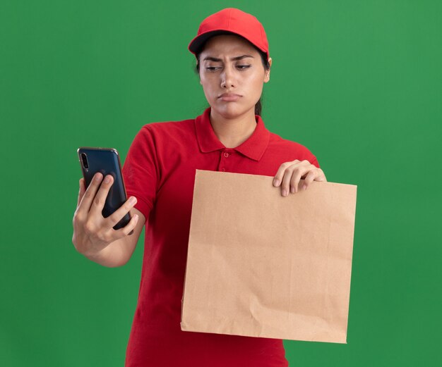 Denken junges Liefermädchen, das Uniform und Kappe trägt, die Papiernahrungsmittelpaket hält und Telefon in ihrer Hand lokalisiert auf grüner Wand betrachtet