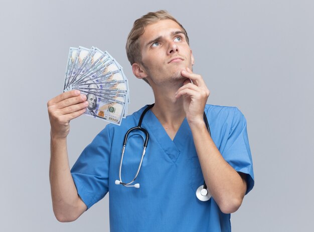Denken nachschlagen junger männlicher Arzt, der Arztuniform mit Stethoskop hält, das Bargeld hält Kinn lokalisiert auf weißer Wand hält