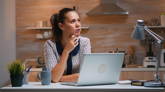 Denken Sie an das nächste Projekt, während Sie von zu Hause aus arbeiten, und lesen Sie die Aufgaben mit dem Laptop in der Küche. Beschäftigter, fokussierter Mitarbeiter, der moderne drahtlose Netzwerktechnologie verwendet, macht Überstunden für den Job