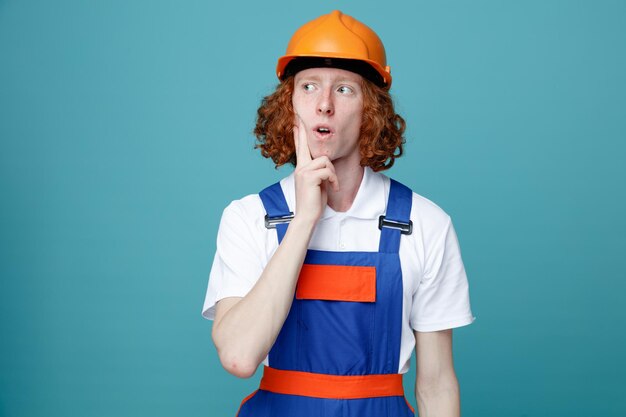 Denken Sie daran, die Finger auf die Wange zu legen. Junger Baumeister in Uniform isoliert auf blauem Hintergrund