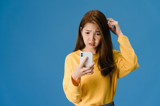 Denken träumende junge asiatische Dame, die Telefon mit positivem Ausdruck verwendet, gekleidet in lässiger Kleidung, die Glück fühlt und lokalisiert auf blauem Hintergrund steht. Glückliche entzückende frohe Frau freut sich über Erfolg.
