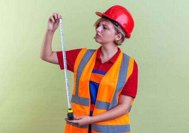 Denkende junge Baumeisterin in Uniform, die Maßband isoliert auf olivgrüner Wand ausstreckt