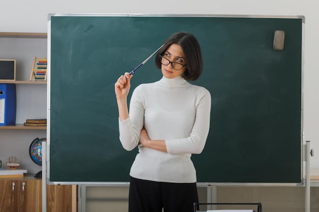 Denkende junge Lehrerin mit Brille, die vor der Tafel steht und den Zeiger auf dem Kopf im Klassenzimmer hält