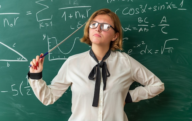 Kostenloses Foto denkende junge lehrerin mit brille, die vor der tafel steht und einen zeiger hält, der die hand auf die hüfte im klassenzimmer legt