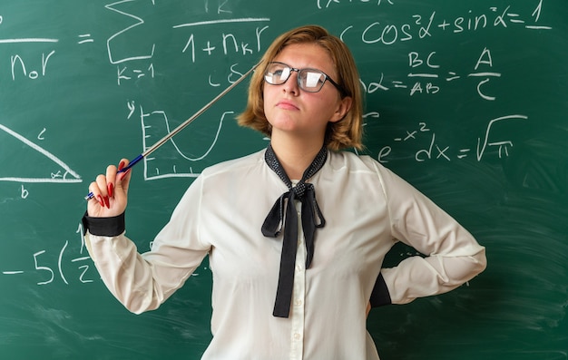 denkende junge Lehrerin mit Brille, die vor der Tafel steht und einen Zeiger hält, der die Hand auf die Hüfte im Klassenzimmer legt