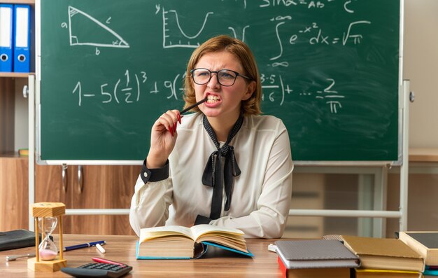 Denkende junge Lehrerin mit Brille sitzt am Tisch mit Schulmaterial, das Bleistift im Klassenzimmer hält