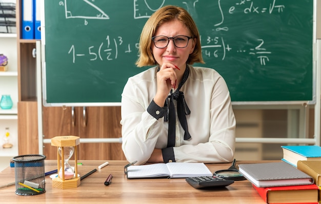 Denkende junge Lehrerin mit Brille sitzt am Tisch mit Schulmaterial, das im Klassenzimmer am Kinn gegriffen hat