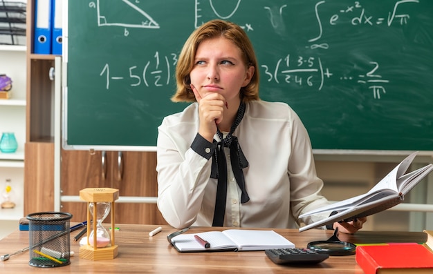 Kostenloses Foto denkende junge lehrerin sitzt am tisch mit schulmaterial, das ein buch hält, das kinn im klassenzimmer gepackt hat