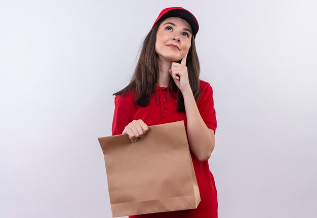 Denkende junge Lieferfrau, die rotes T-Shirt in der roten Kappe hält, die ein Paket auf isolierter weißer Wand hält