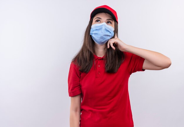 Denkende junge Lieferfrau, die rotes T-Shirt in roter Kappe trägt, trägt Gesichtsmaske auf isolierter weißer Wand