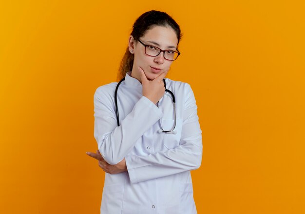 Denkende junge Ärztin, die medizinische Robe und Stethoskop mit Brille trägt, packte Kinn lokalisiert auf orange Wand