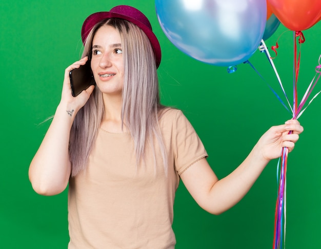 Denkende junge schöne Mädchen mit Partyhut und Hosenträgern, die Luftballons halten, spricht am Telefon isoliert auf grüner Wand