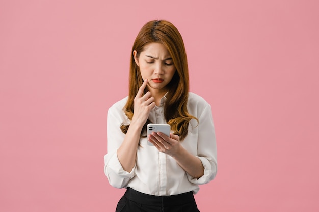 Denkende träumende junge Asiatin mit Telefon mit positivem Ausdruck, gekleidet in Freizeitkleidung, die sich glücklich fühlt und einzeln auf rosafarbenem Hintergrund steht.