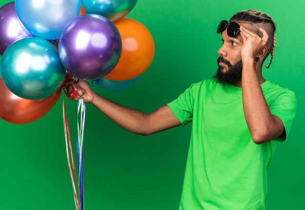 Kostenloses Foto denkender junger afroamerikanischer mann mit brille, der ballons hält und betrachtet