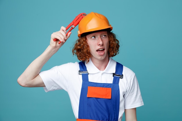 Denkender junger Baumeister in Uniform mit Gasschlüssel auf dem Kopf isoliert auf blauem Hintergrund