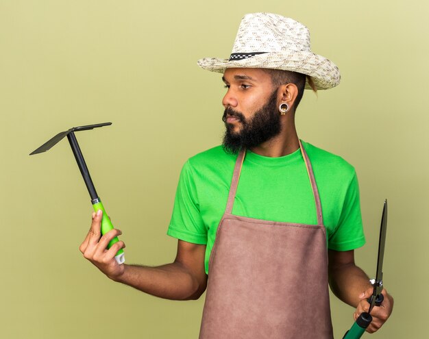 Denkender junger Gärtner afroamerikanischer Mann mit Gartenhut, der Schermaschinen mit Hackeharke hält