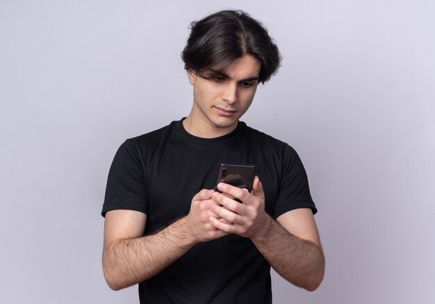 Denkender junger gutaussehender Kerl, der schwarzes T-Shirt hält hält und Telefon lokalisiert auf weißer Wand betrachtet